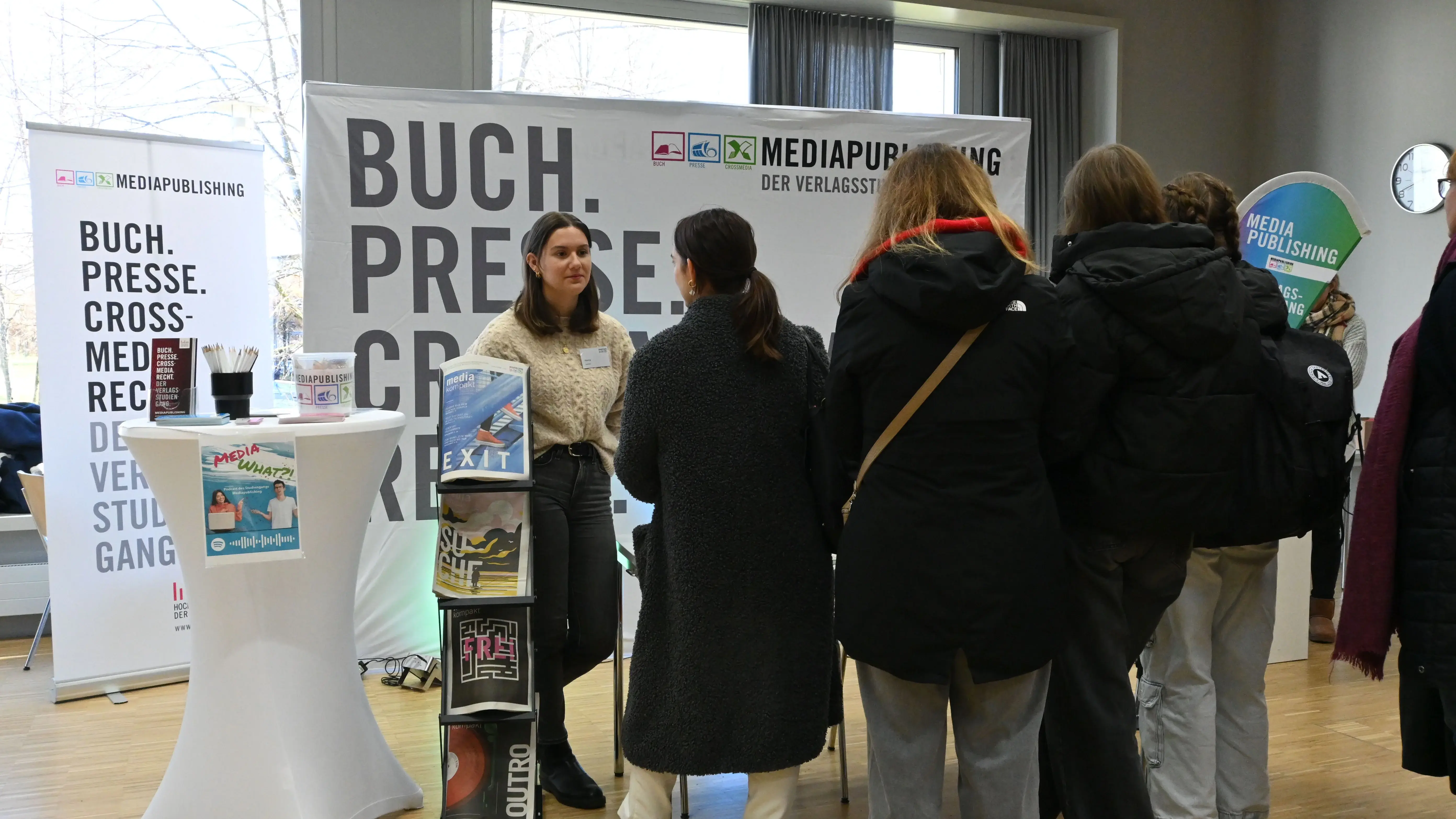 Auf dem Foto sind man den Präsentationsstand des Studiengangs MEdiapublishing auf dem Studieninfotag 2025. Im Vordergrund stehen Personen, die mit Studierenden vom Infostand im Gespräch sind.